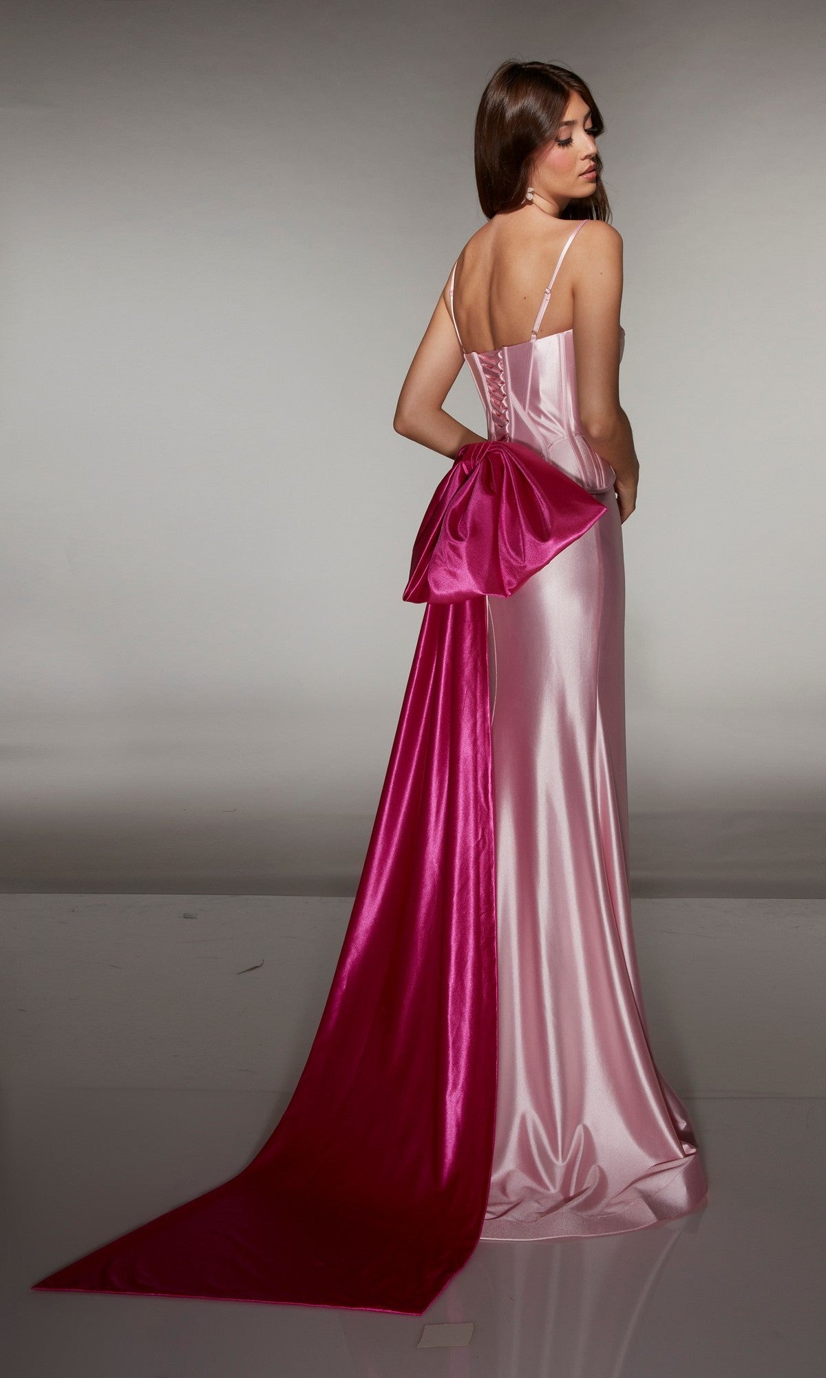 A woman models the Alyce 61983 long prom dress: a shiny light pink satin gown with thin straps, a fitted corset bodice, and a large magenta bow with detachable sweep train, set against a plain background.